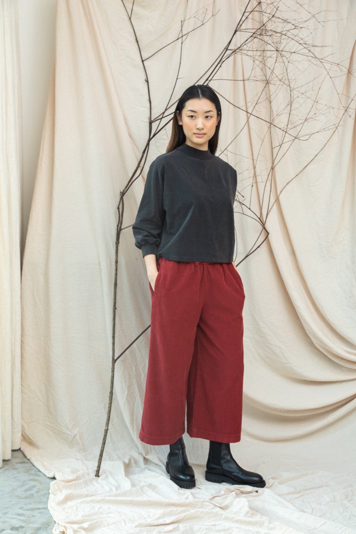 Person wearing a dark sweater and red pants standing against a beige curtain with branches.