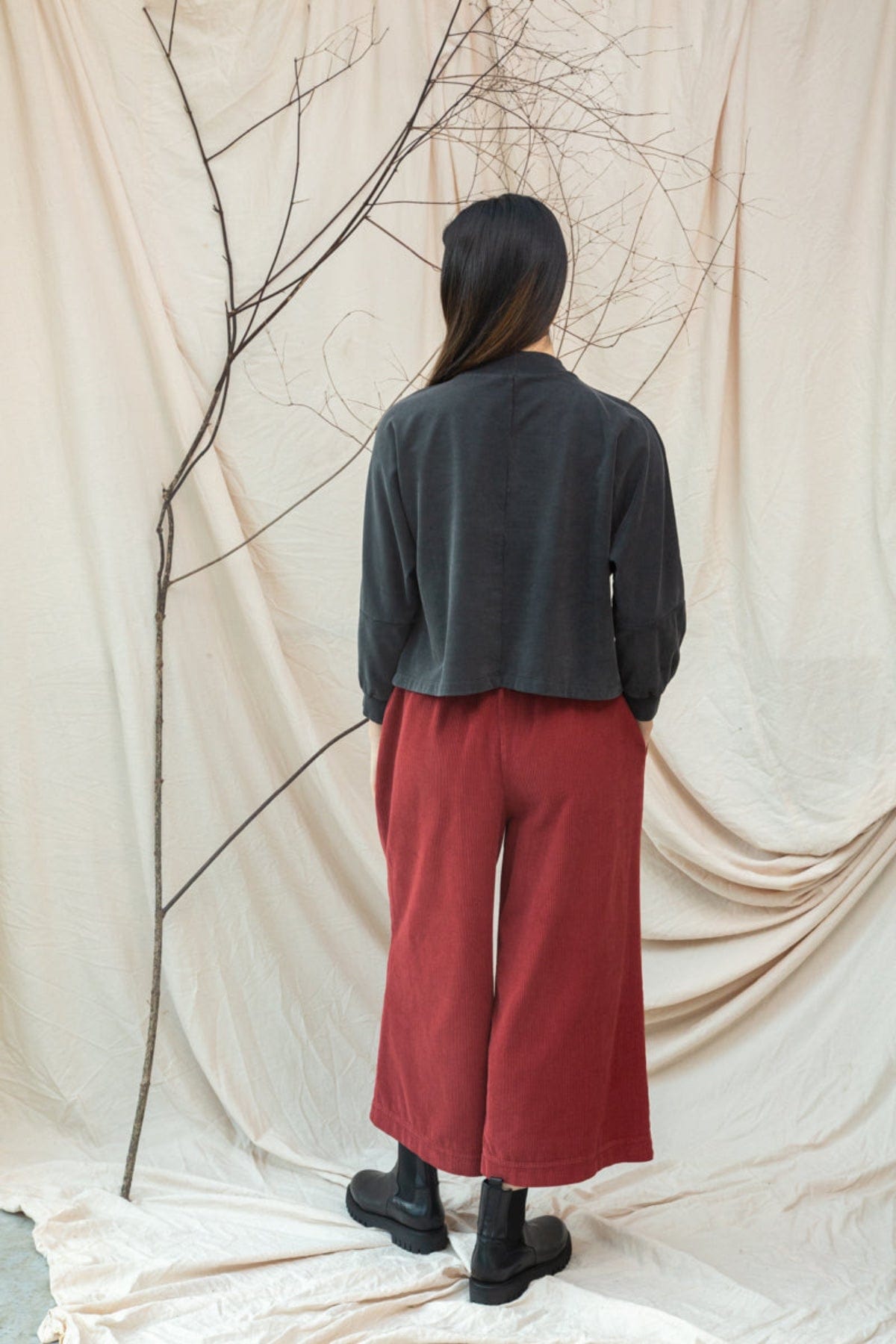 Person wearing a dark shirt and red pants standing against a draped beige curtain with a branch.