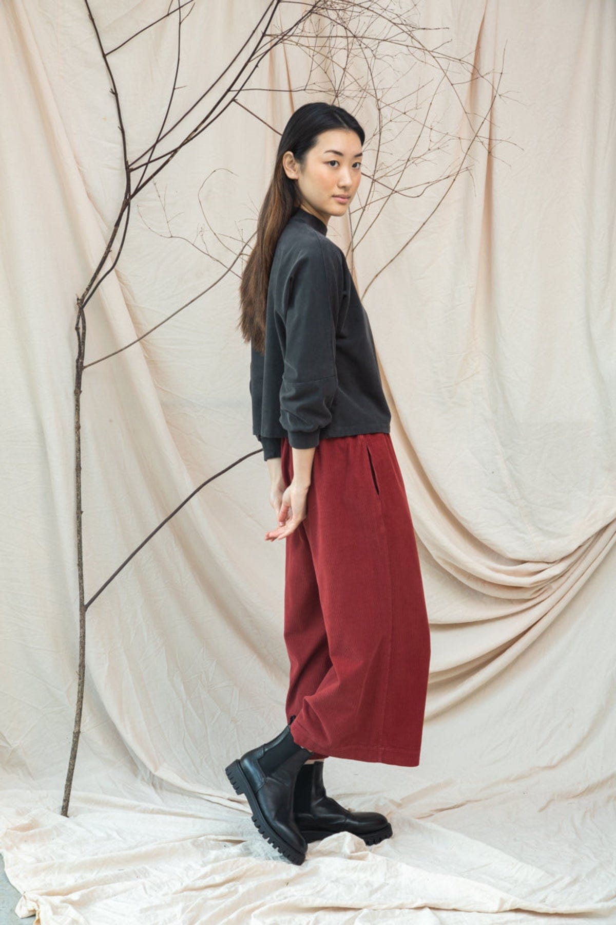 Person wearing a black top and red pants standing against a beige curtain with bare branches.