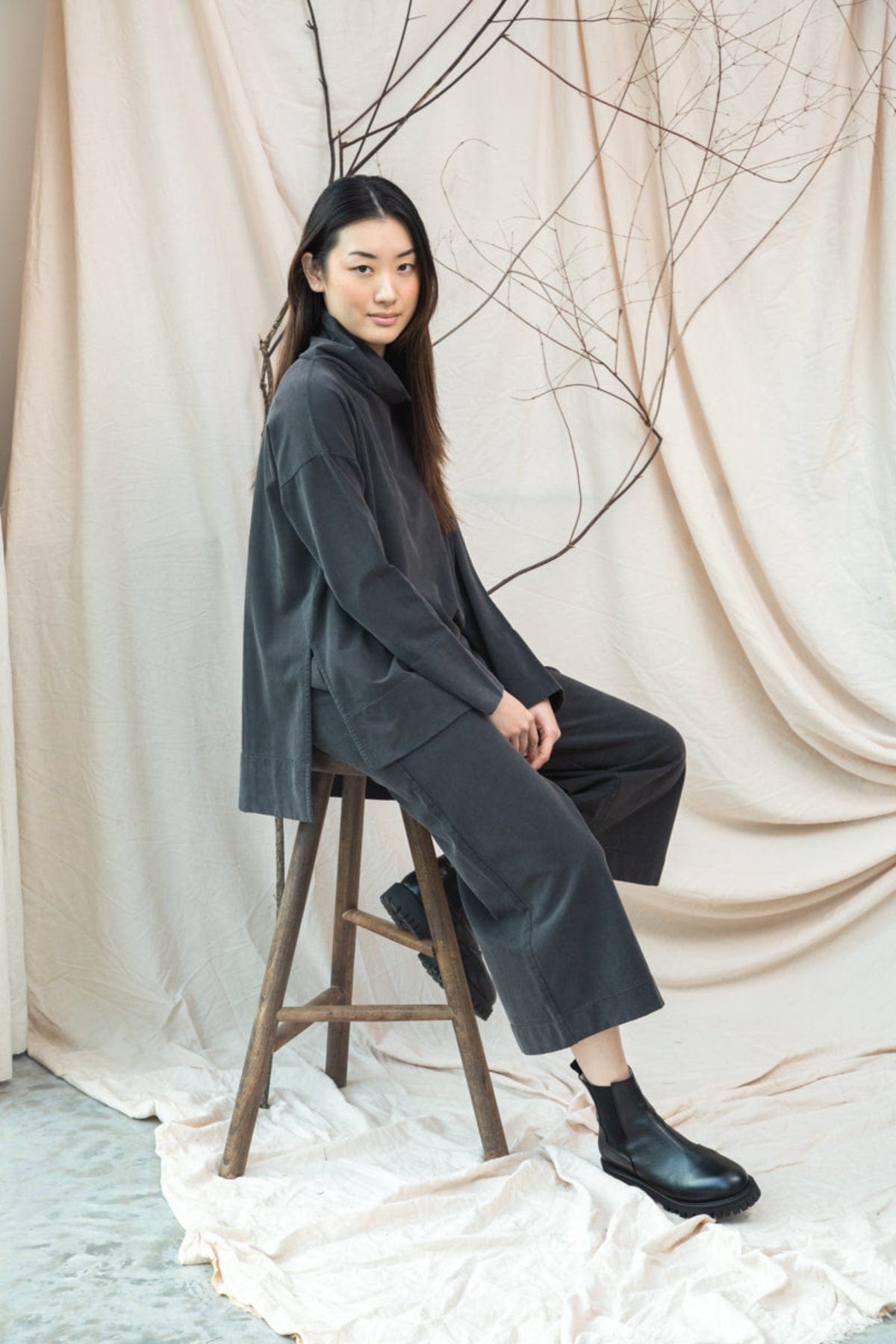 Woman sitting on a stool against a beige curtain with bare branches.
